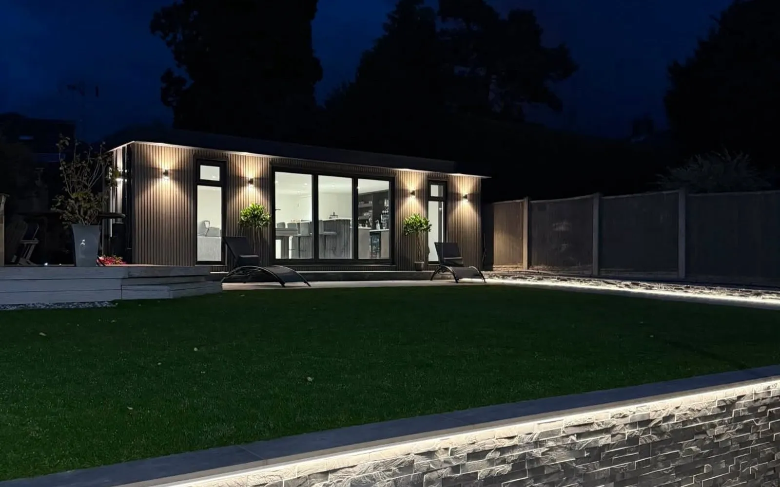 Wide evening view of a bespoke garden entertainment room with warm interior lighting